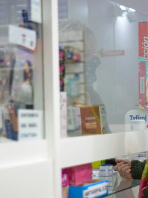 A woman with a mask speaks to a pharmacist behind a counter in a pharmacy.