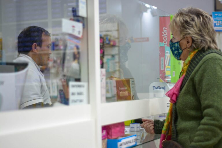 A woman with a mask speaks to a pharmacist behind a counter in a pharmacy.
