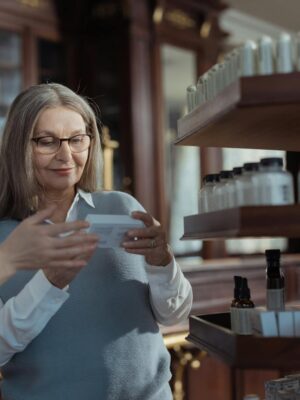 Elderly woman receiving assistance from a pharmacist in a pharmacy with product shelves.